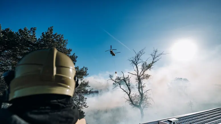 Brandweer blust brand terwijl in de lucht een Chinook-blushelikopter water dropt. 