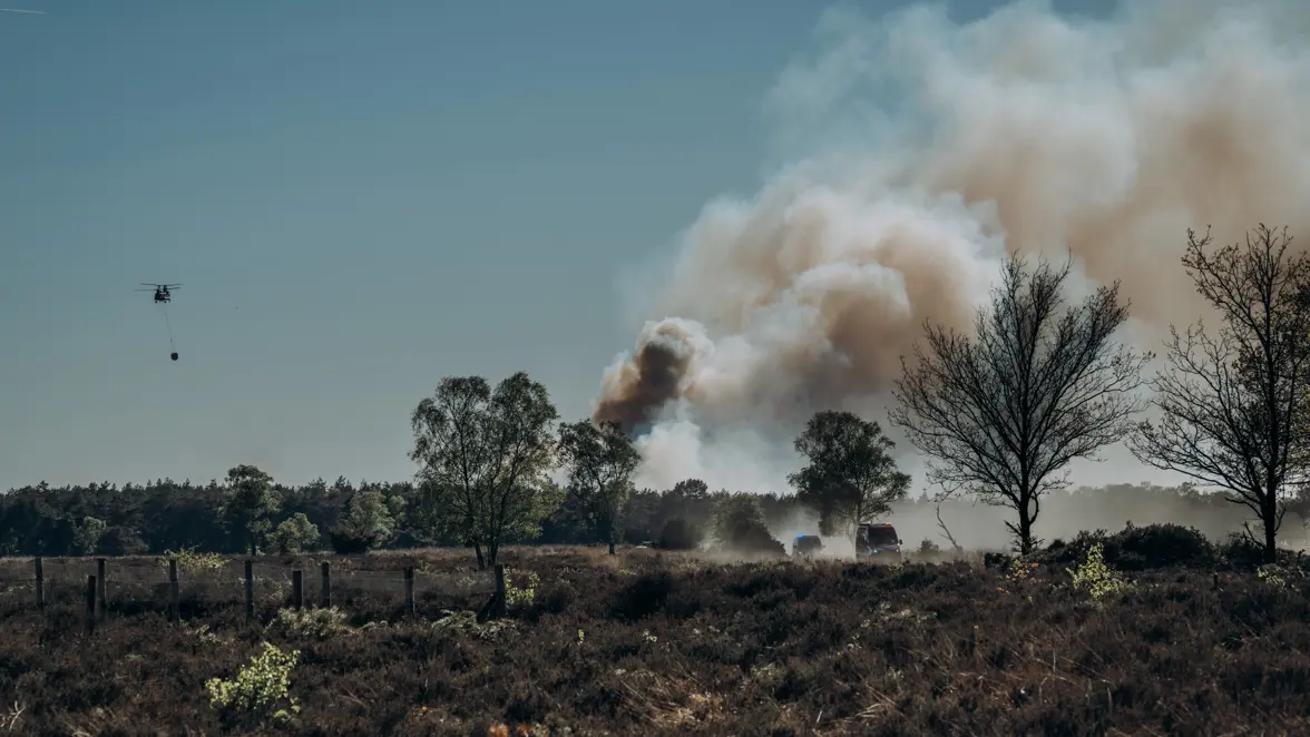 Een Chinook-transporthelikopter vliegt in de verte boven een bos, terwijl rookwolken opstijgen. 