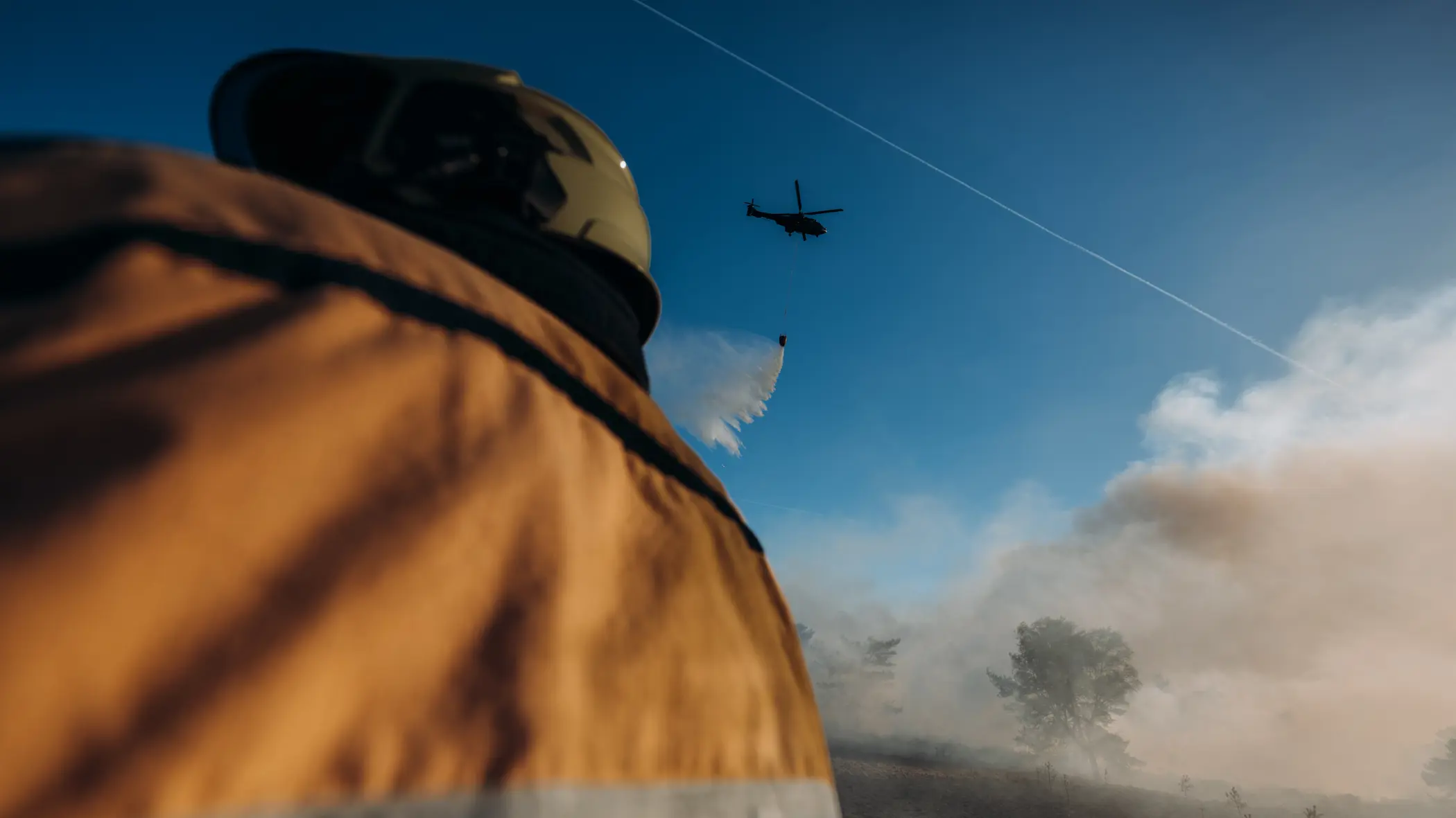 Een brandweerman kijkt omhoog en ziet hoe een helikopter bluswater loost op een brandhaard. 