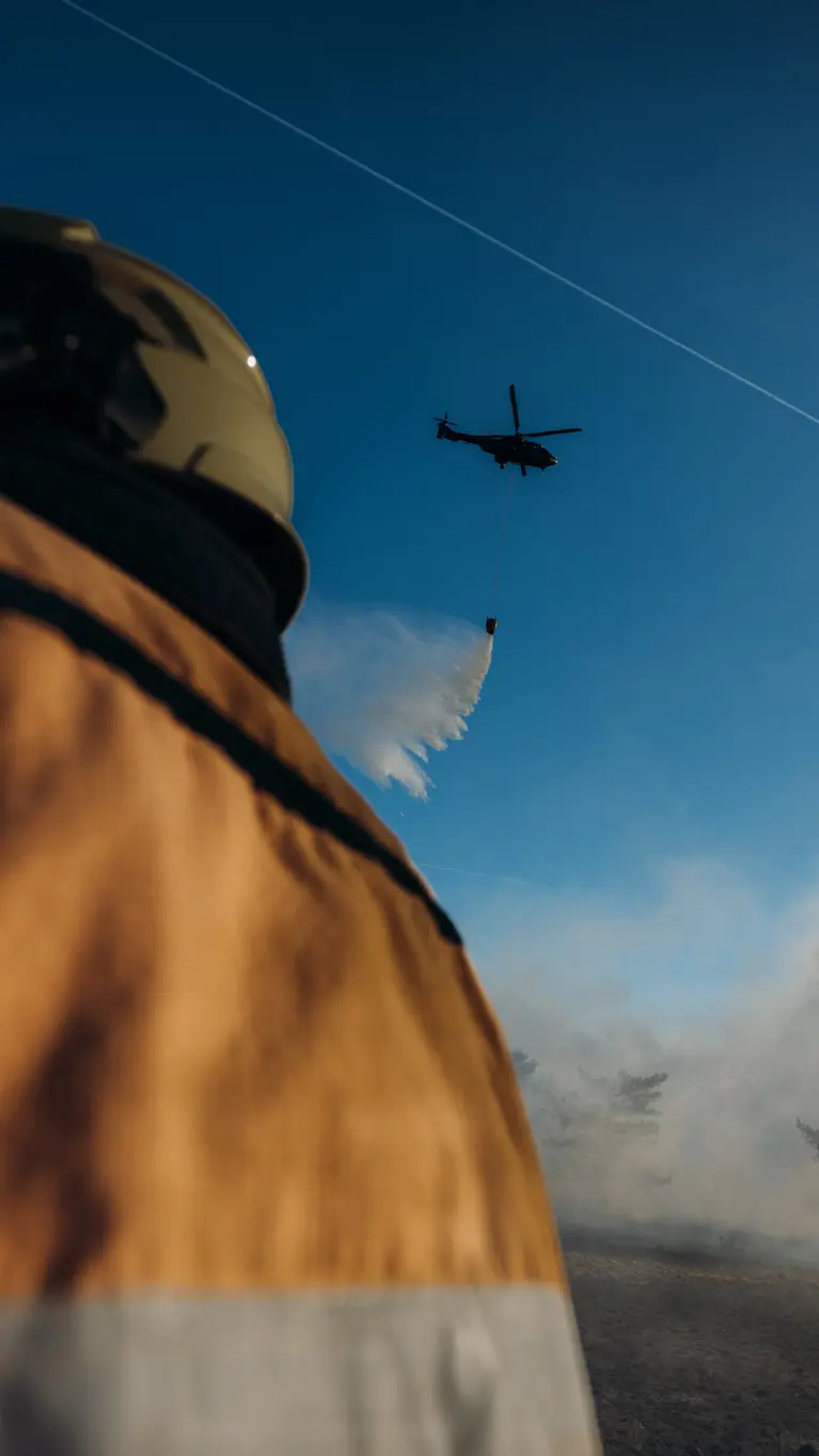 Een brandweerman kijkt omhoog en ziet hoe een helikopter bluswater loost op een brandhaard. 