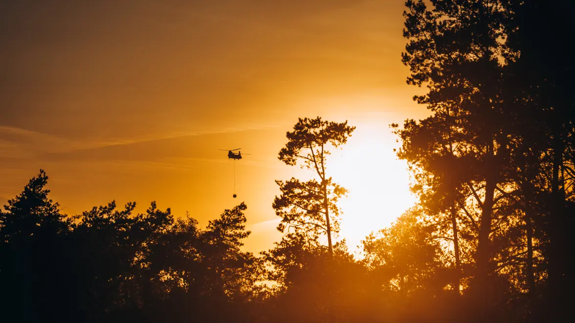 Een Chinook-transporthelikopter vliegt boven een bos de zonsondergang tegemoet. Onder de heli hangt een waterzak om branden te blussen.