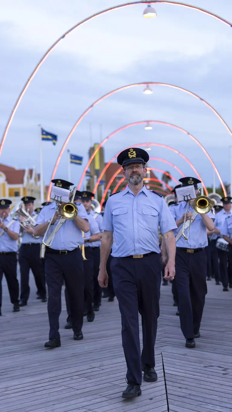 Orkest met blazers vooraan loopt achter man aan brug met lampjes over. 