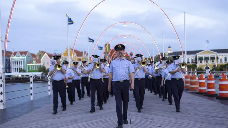Orkest met blazers vooraan loopt achter man aan brug met lampjes over. 
