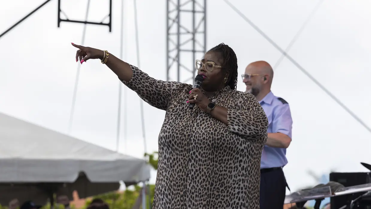 Donkere bebrilde zangeres zingt op podium en wijst naar publiek. 