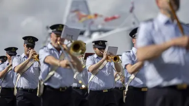Muzikanten van luchtmacht spelen op hun instrumenten met op achtergrond een marineschip. 