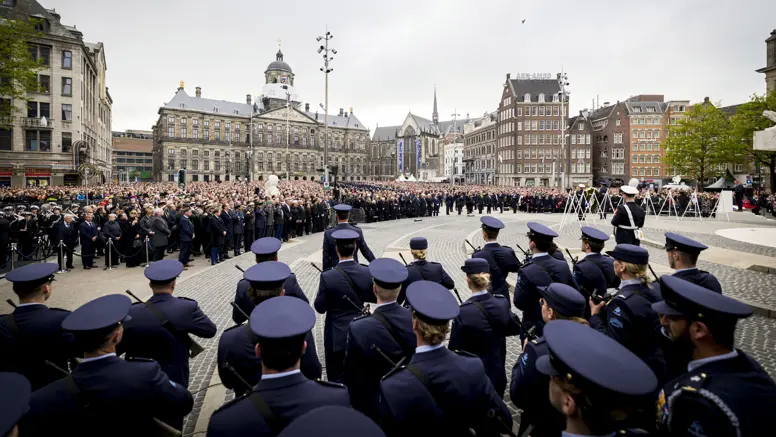 Beeld van de Dam in Amsterdam met de blik pop het Paleis op de Dam, met in de voorgrond op de rug gekeken opgestelde luchtmachtmilitairen. Daartegenover staan vele honderden genodigden en bezoekers van de herdenking.