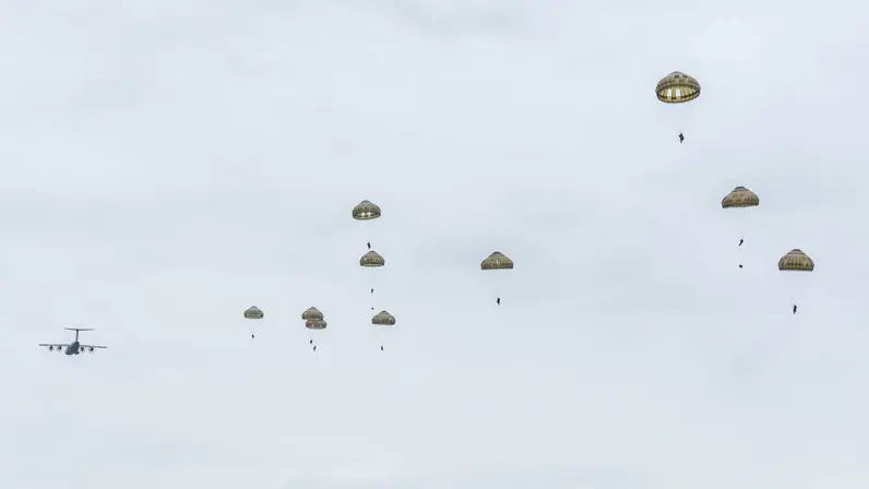 Tien parachutisten in de lucht met op de achtergrond een wegvliegend transportvliegtuig.