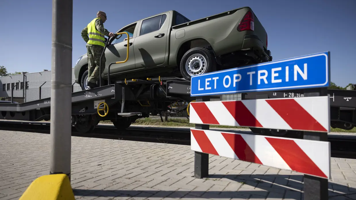 Een pick-up truck staat op een goederenwagon, ernaast staat een militair in geel hesje. Op de voorgrond staat een rood-wit bord met erboven een blauw bord met de tekst ‘let op trein’. 