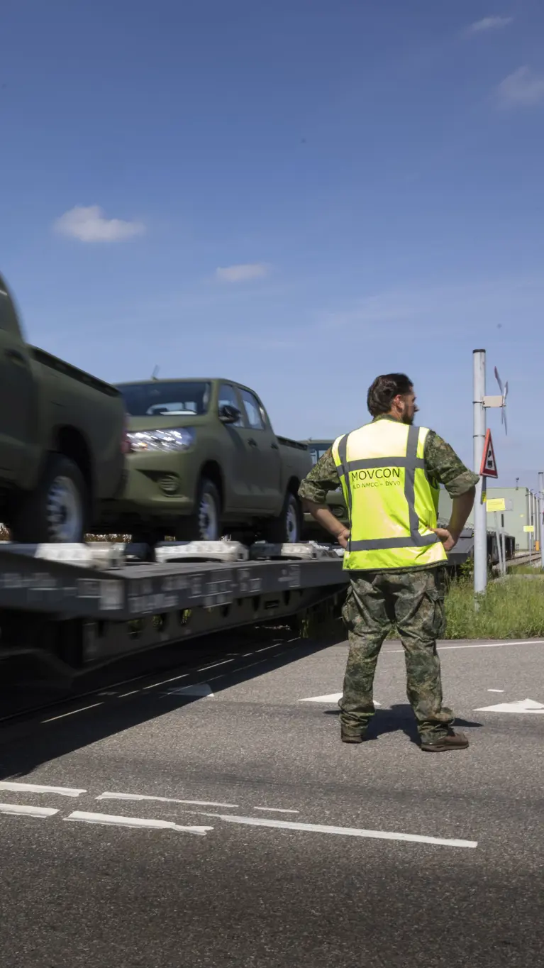 Een militair in geel hesje staat bij een voorbijrijdende trein met daarop voertuigen. 