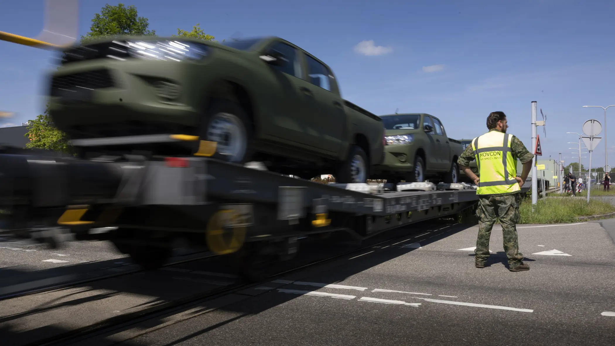 Een militair in geel hesje staat bij een voorbijrijdende trein met daarop voertuigen. 