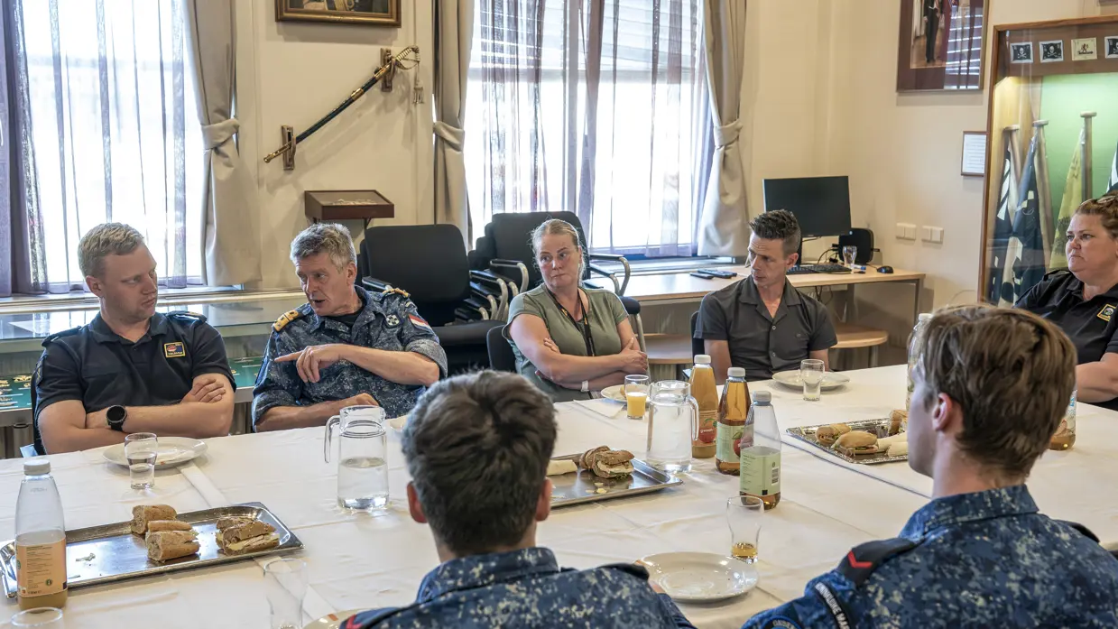 5 marinemensen en 2 mensen in burgerkleding overleggen met elkaar aan tafel tijdens een lunch die op tafel staat.