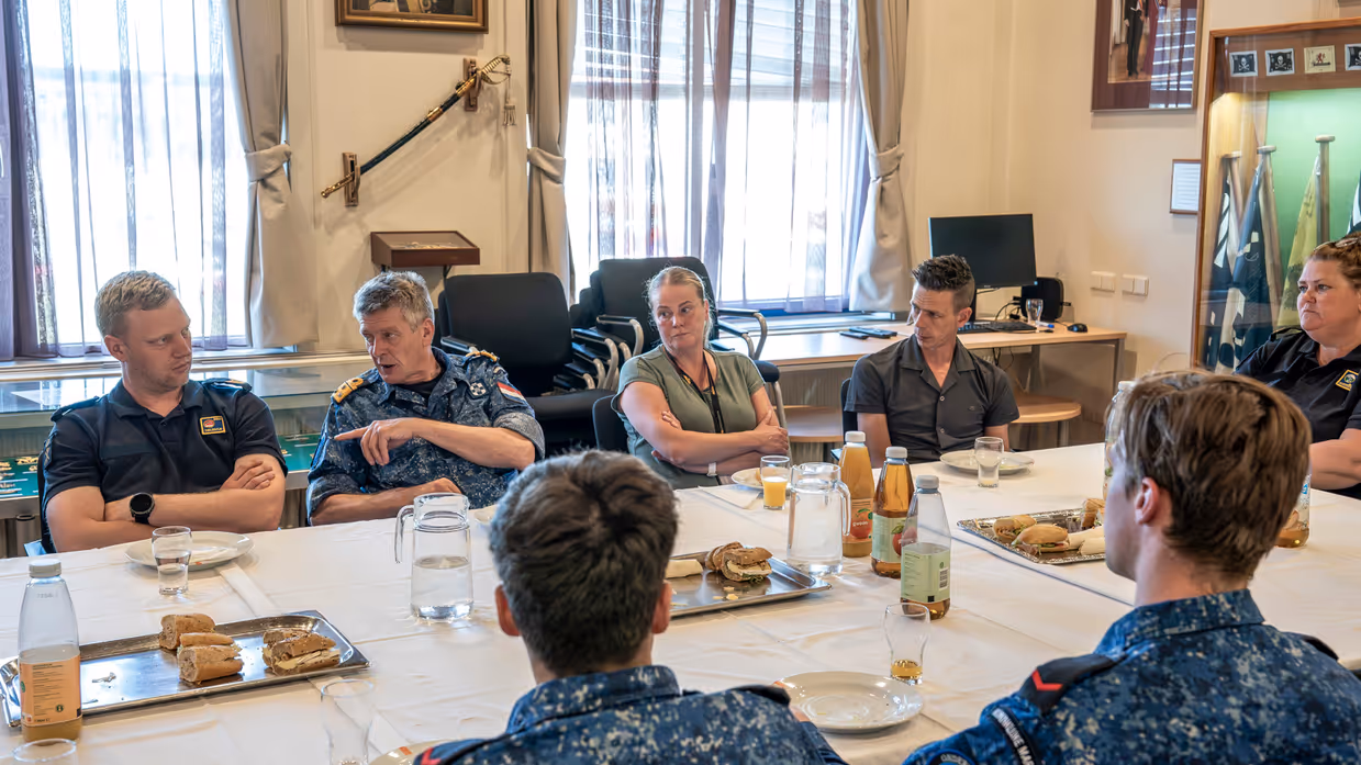 5 marinemensen en 2 mensen in burgerkleding overleggen met elkaar aan tafel tijdens een lunch die op tafel staat.