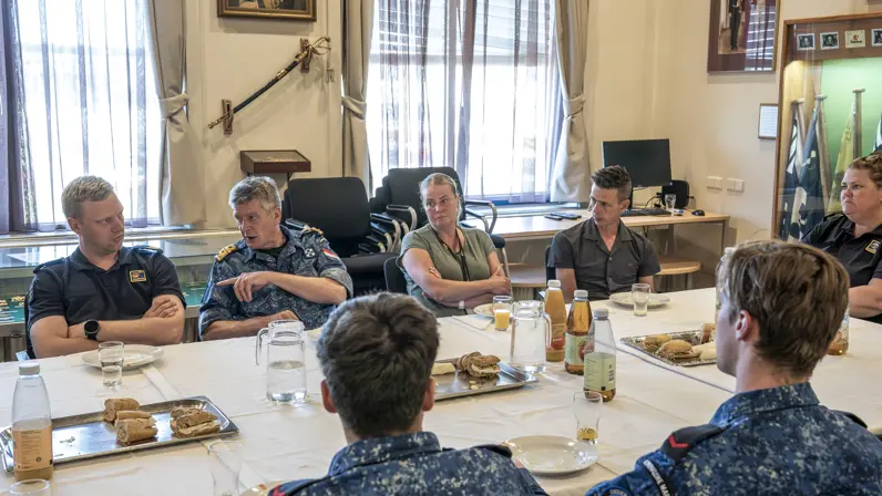 5 marinemensen en 2 mensen in burgerkleding overleggen met elkaar aan tafel tijdens een lunch die op tafel staat.