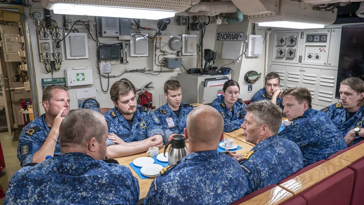 10 marinemensen praten met elkaar aan een tafel in een onderzeeboot.&nbsp;
