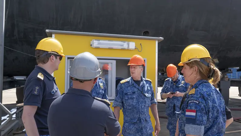  6 marinemensen met bouwhelm op, van wie 1 onzichtbaar is, praten met elkaar voor een geel hokje.