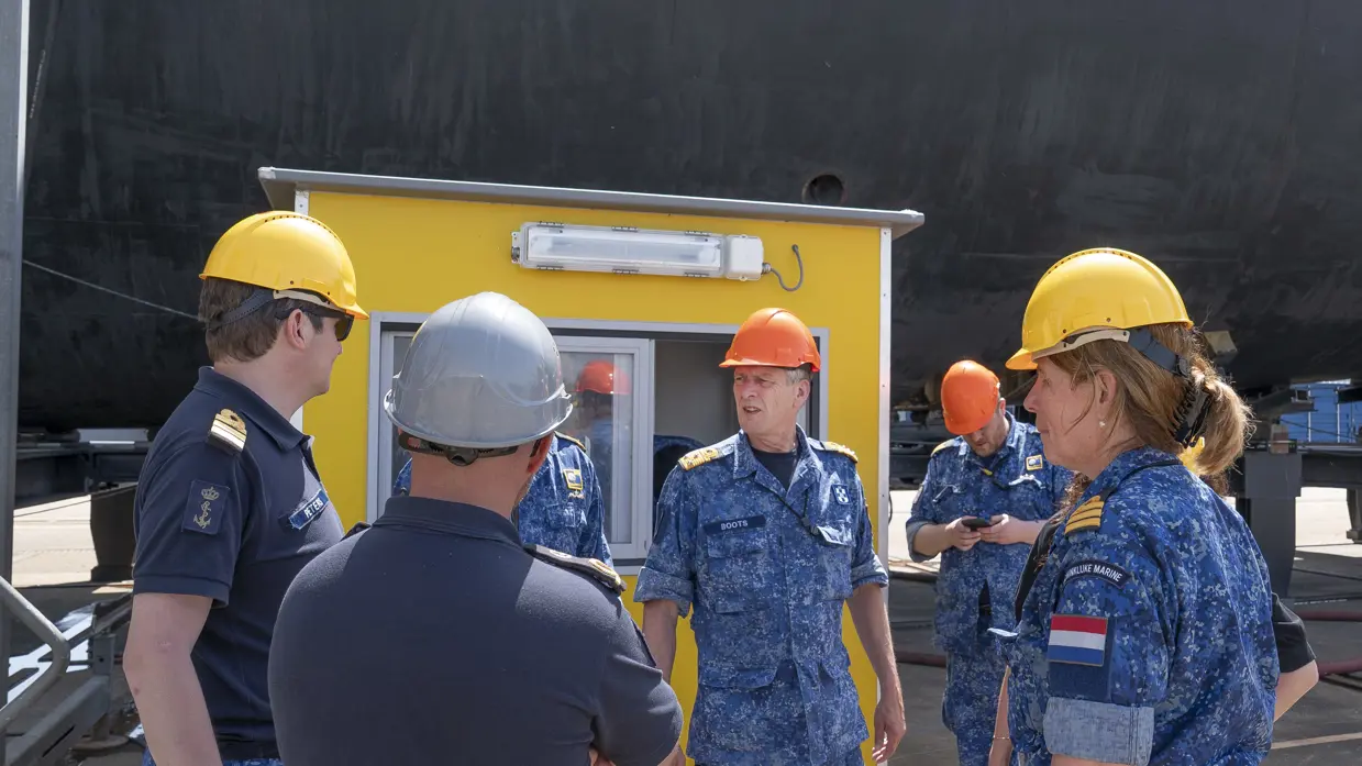  6 marinemensen met bouwhelm op, van wie 1 onzichtbaar is, praten met elkaar voor een geel hokje.