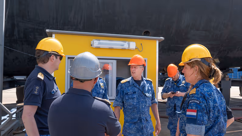  6 marinemensen met bouwhelm op, van wie 1 onzichtbaar is, praten met elkaar voor een geel hokje.
