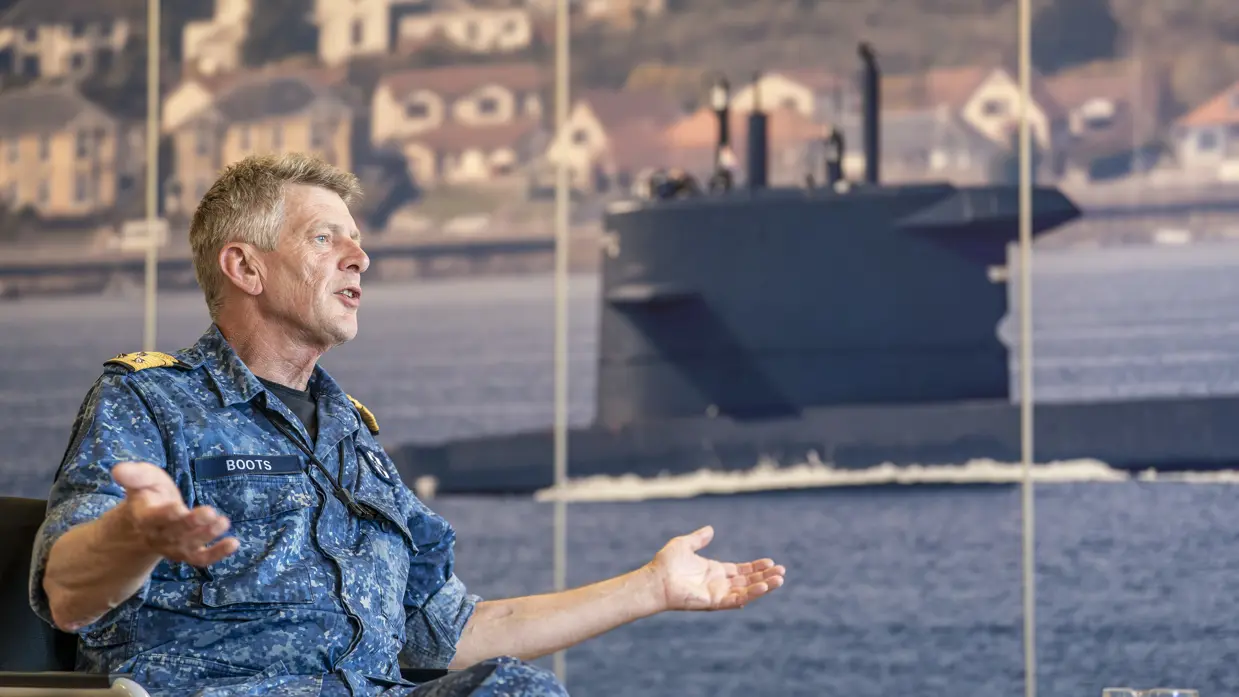 Een viceadmiraal praat met gespreide armen en open handen met op de achtergrond een foto van een onderzeeboot.