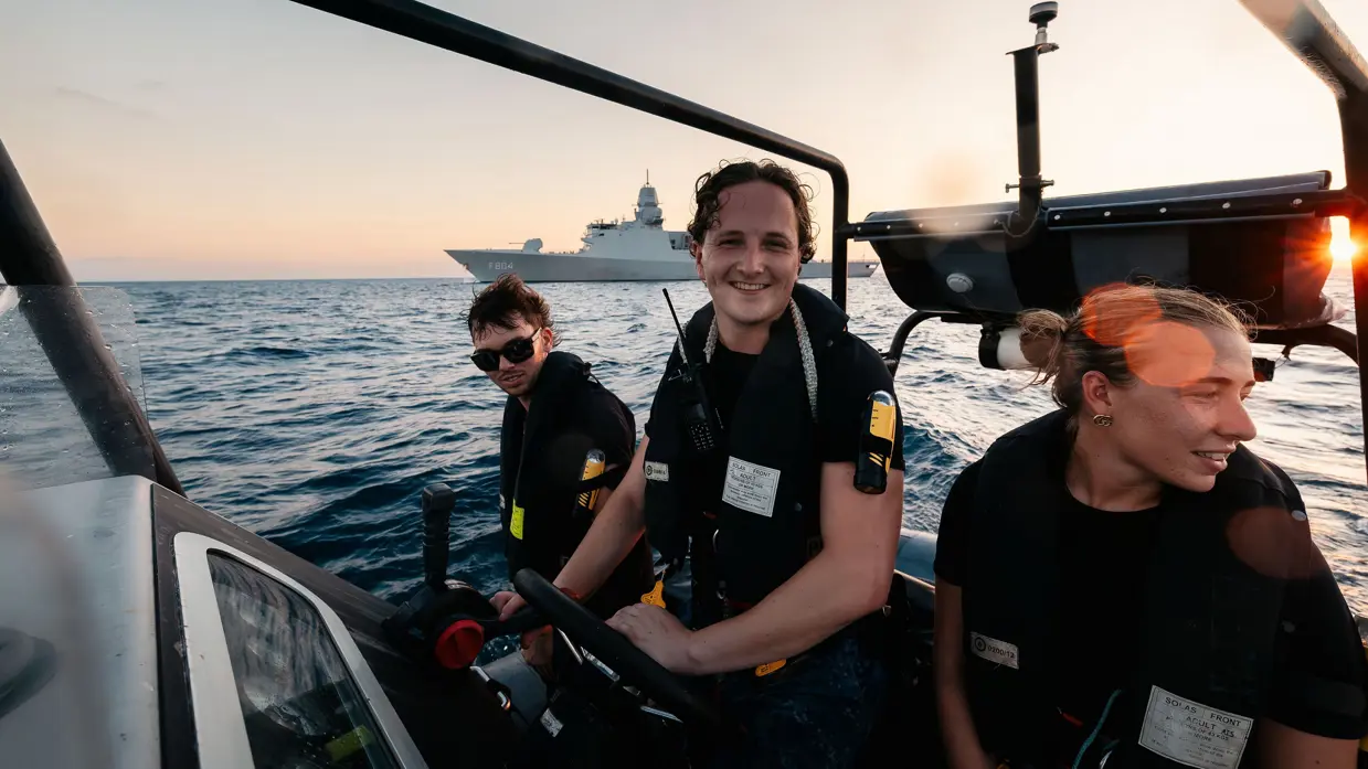 Drie militairen op een boot, met een schip in de achtergrond.
