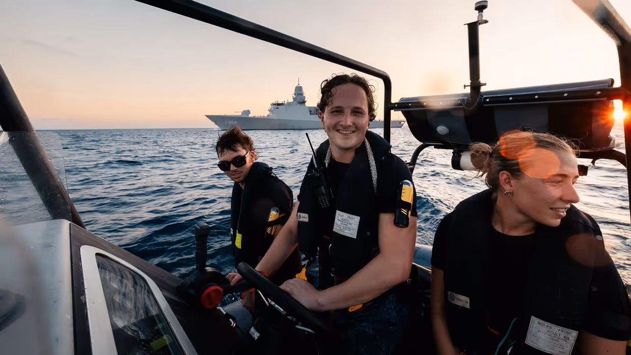Drie militairen op een boot, met een schip in de achtergrond.