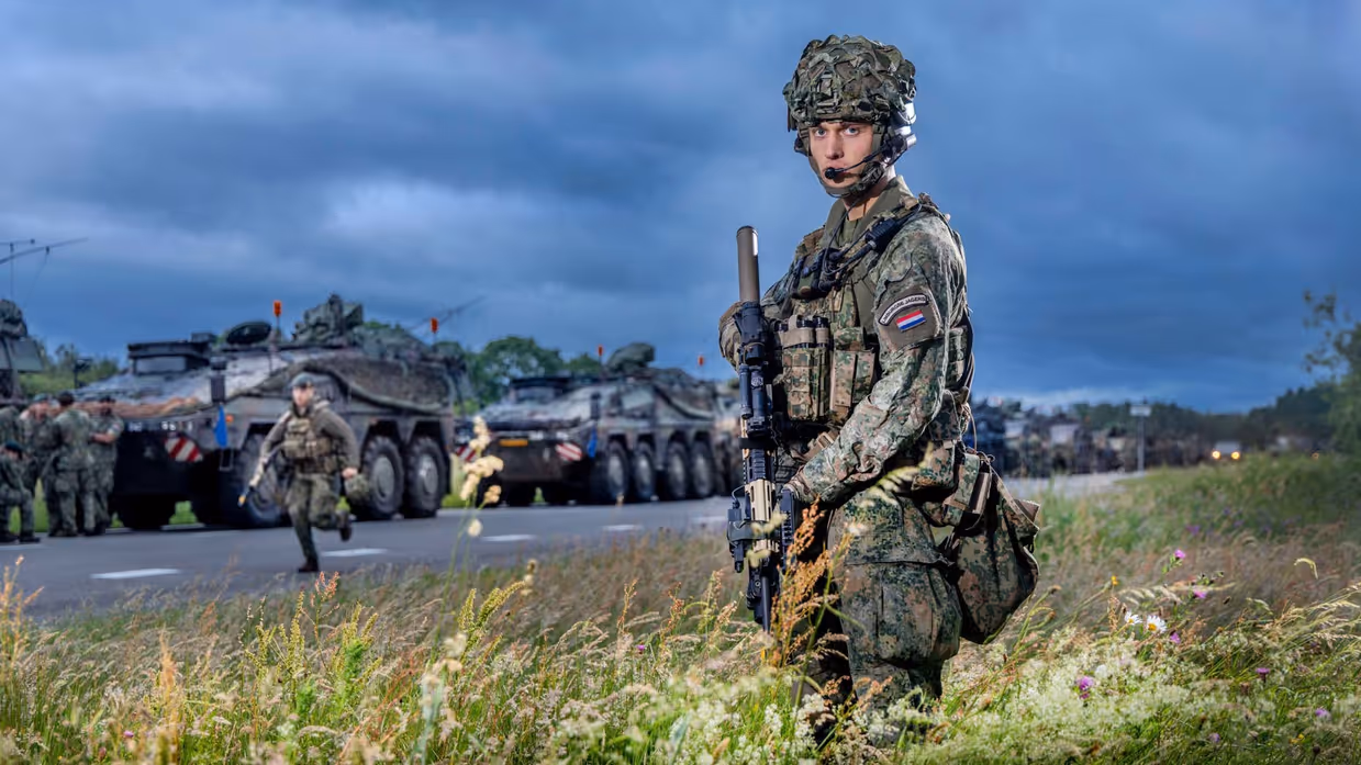 Een militair poseert terwijl de colonne aan militaire trucks achter hem in een rij aan de weg staan.