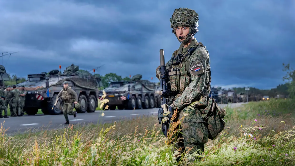 Een militair poseert terwijl de colonne aan militaire trucks achter hem in een rij aan de weg staan.