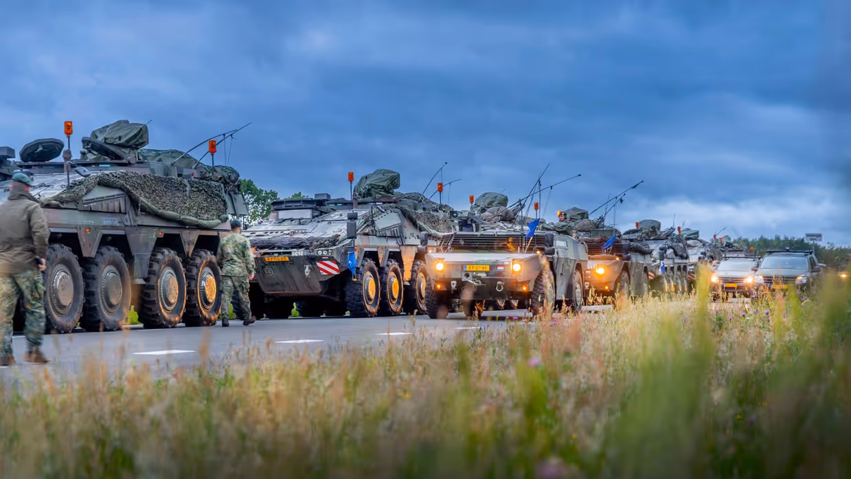 Militaire trucks staan stil op de weg. Militairen lopen rond terwijl burgerauto's in de achtergrond staan.