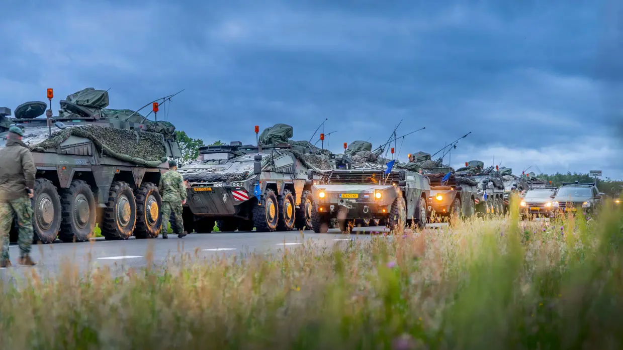 Militaire trucks staan stil op de weg. Militairen lopen rond terwijl burgerauto's in de achtergrond staan.