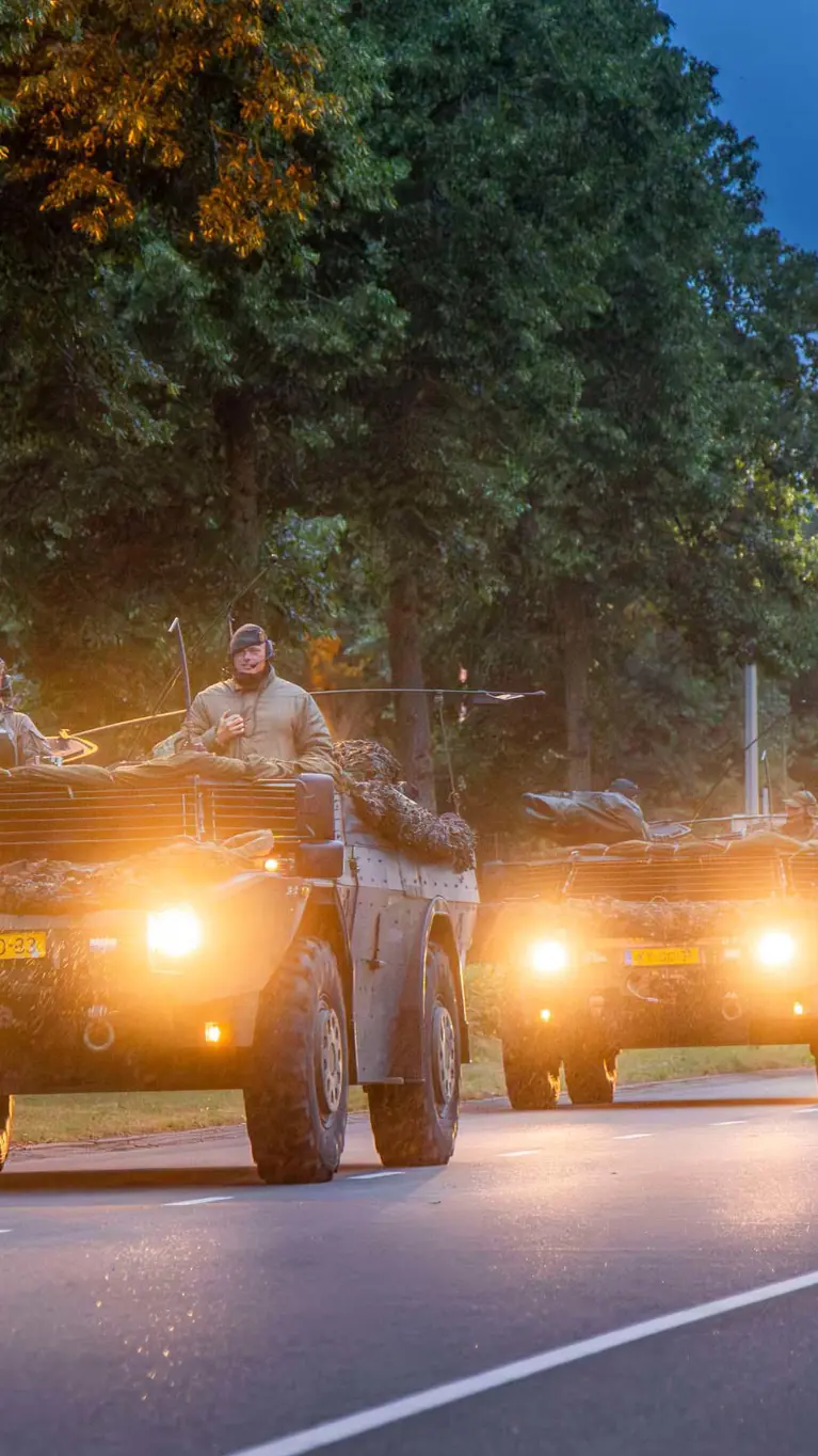 Militaire voertuigen rijden ’s avonds in colonne over een weg.