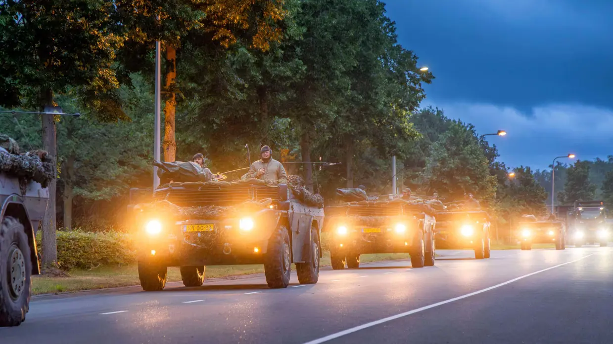 Een colonne van vier militaire wagens rijdt over de weg met koplampen aan.