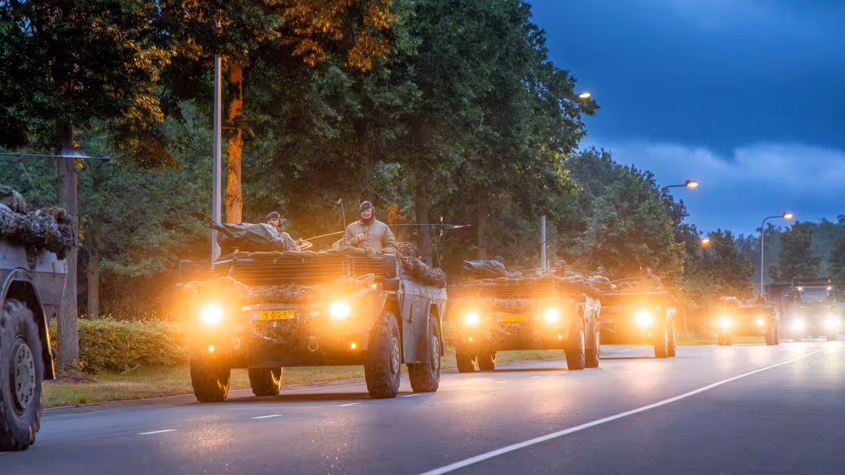 Een colonne van vier militaire wagens rijdt over de weg met koplampen aan.