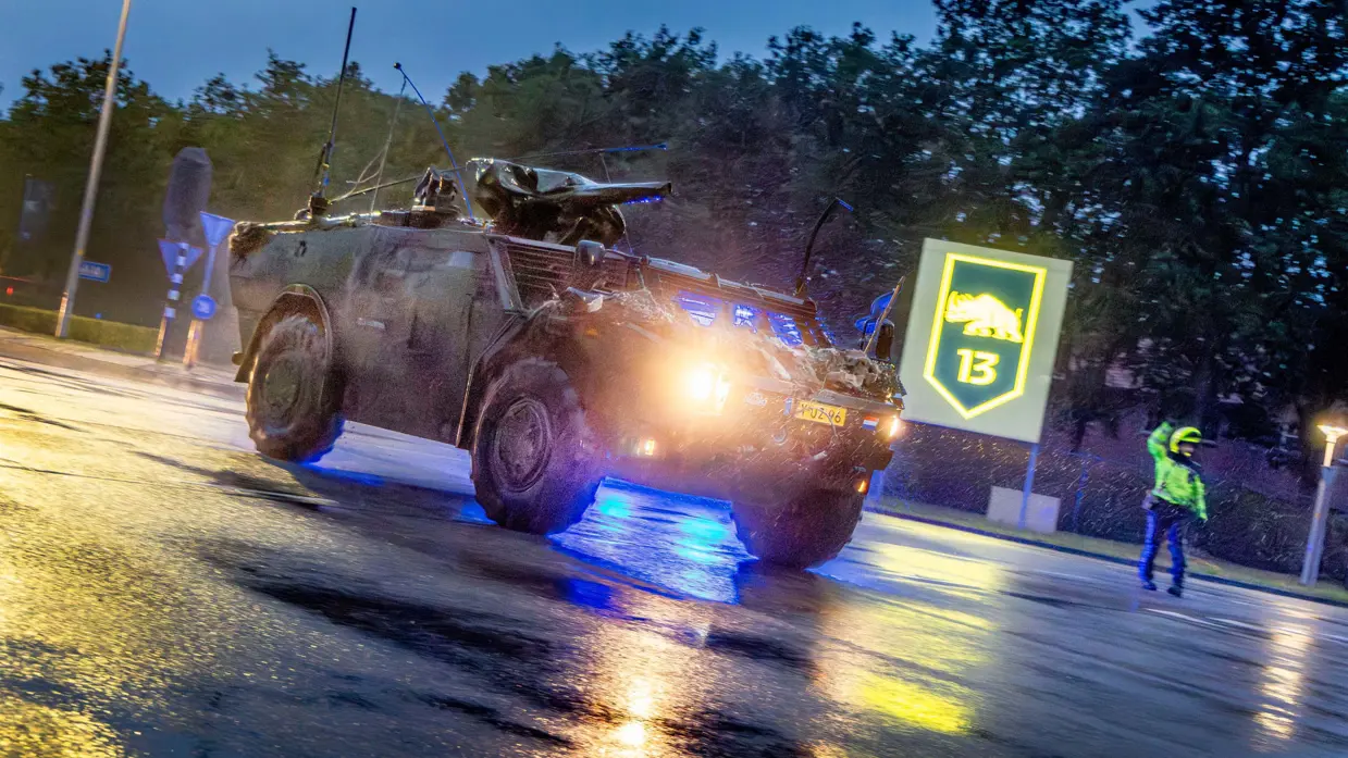 Een militaire truck rijdt over een weg met in de achtergrond een agent die verkeer reguleert.