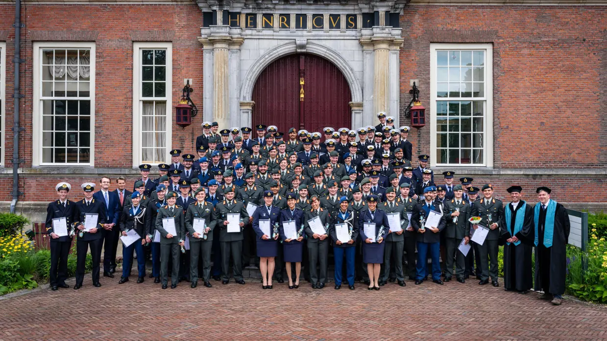 Grote groep militairen, afkomstig van alle krijgsmachtdelen, met in hun handen een diploma. Ze staan voor een grote kasteeldeur van een gebouw met rode bakstenen en hoge ramen.