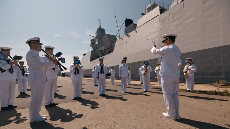Marinemensen in DT staan met instrumenten op een kade bij een groot marineschip. 