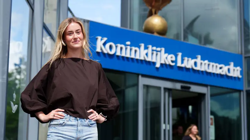  Floor staat voor de ingang van het hoofdkwartier van de Koninklijke Luchtmacht. Op de achtergrond zijn het logo en de ingang van het gebouw met de tekst 'Koninklijke Luchtmacht' zichtbaar.