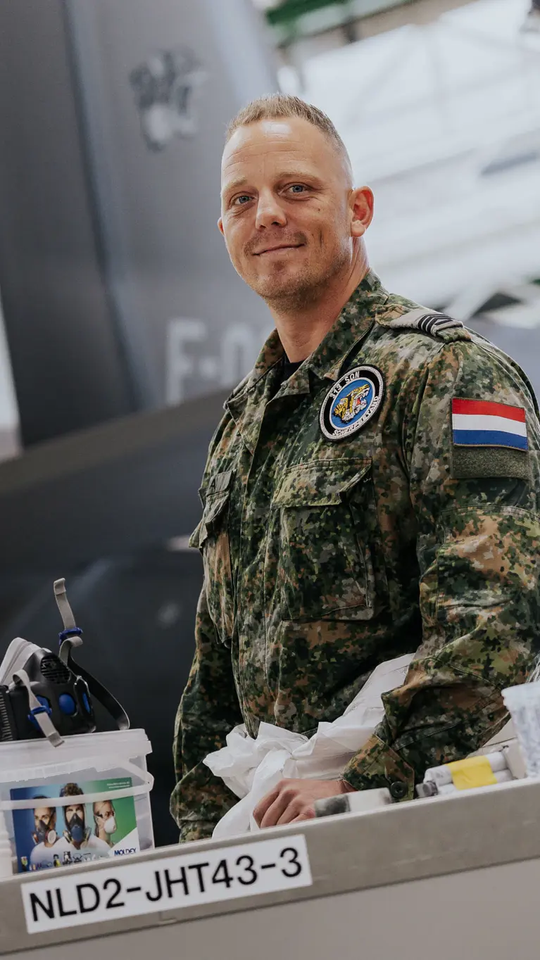 Sergeant 1 Glenn Afman poseert achter een werktafel met kleine spullen erop in een hangaar. Op de achtergrond staat een F-35.