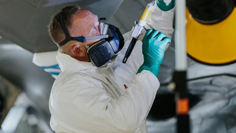  Glenn Afman in witte overal met groene latex handschoenen en een grote zwarte mondkap met luchtfilters op. Met een spuit richt hij op een onderdeel van de F-35.