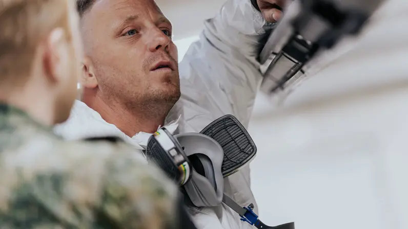 Glenn Afman leunend op een onderdeel van de F-35 terwijl hij luistert naar een collega naast hem in groen vlekkenpak. Glenn draag een witte overal, zijn zwarte mondkap is af en hangt op zijn borst.