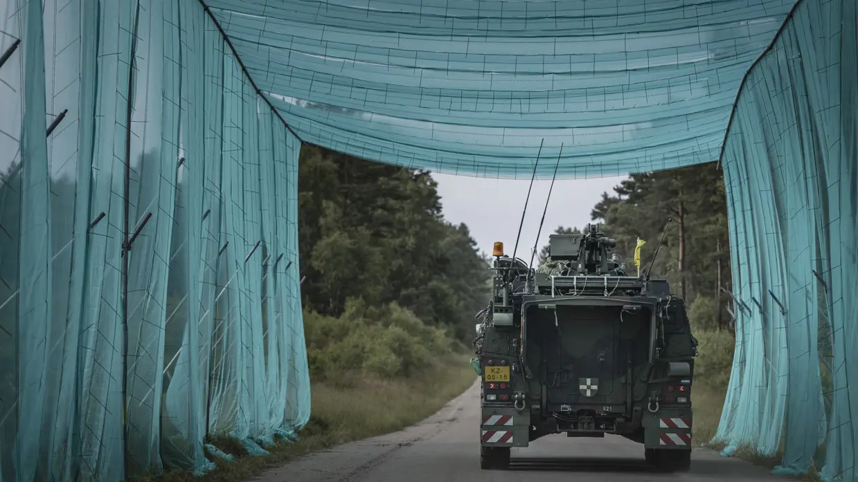 Een militair voertuig rijdt onder een lange strook blauwe camouflagenetten door op een bosweg tijdens oefening Fighter Lion.