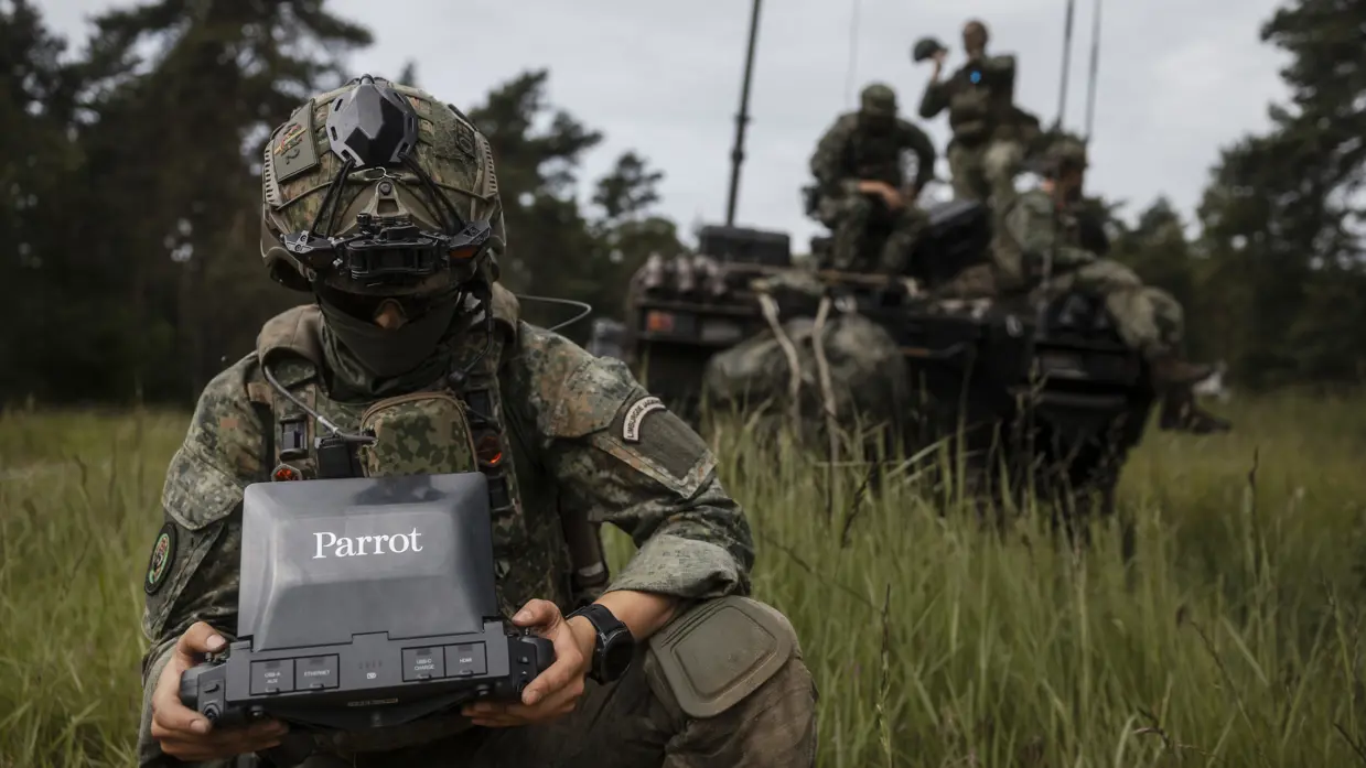 Een militair van 43 Gemechaniseerde Brigade bedient een dronebesturingssysteem terwijl achter hem collega's op een militair voertuig zitten.