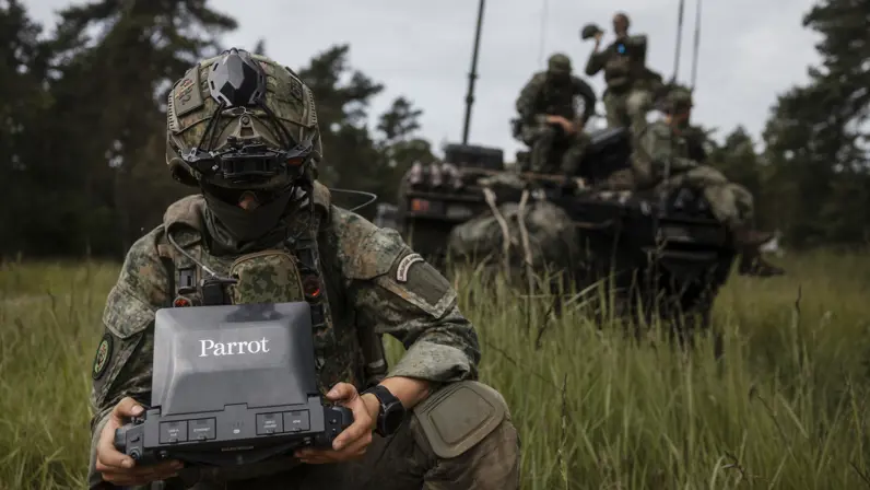 Een militair van 43 Gemechaniseerde Brigade bedient een dronebesturingssysteem terwijl achter hem collega's op een militair voertuig zitten.