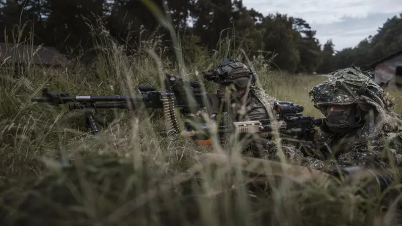 Twee militairen liggen gecamoufleerd in hoog gras tijdens oefening Fighter Lion. Eén bedient een zwaar machinegeweer, terwijl de ander met zijn wapen de omgeving beveiligt. 