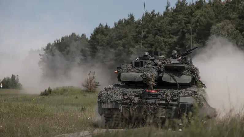 Een Leopard 2A8-gevechtstank rijdt tijdens oefening Fighter Lion over een zandpad. Stof wolkt op terwijl twee militairen vanuit de toren de omgeving observeren.