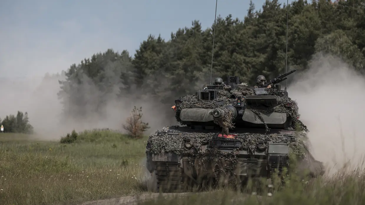Een Leopard 2A8-gevechtstank rijdt tijdens oefening Fighter Lion over een zandpad. Stof wolkt op terwijl twee militairen vanuit de toren de omgeving observeren.