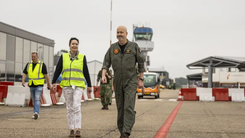 Vrouw in geel hesje loopt naast militair op vliegveld.