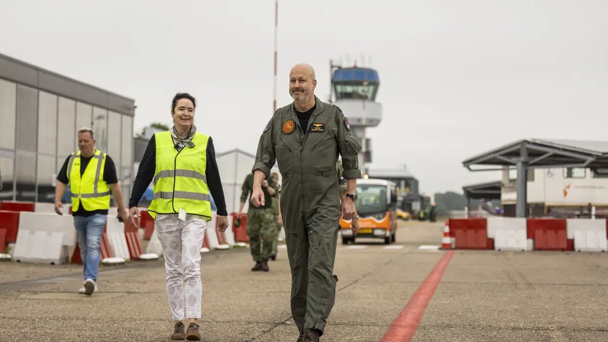 Vrouw in geel hesje loopt naast militair op vliegveld.