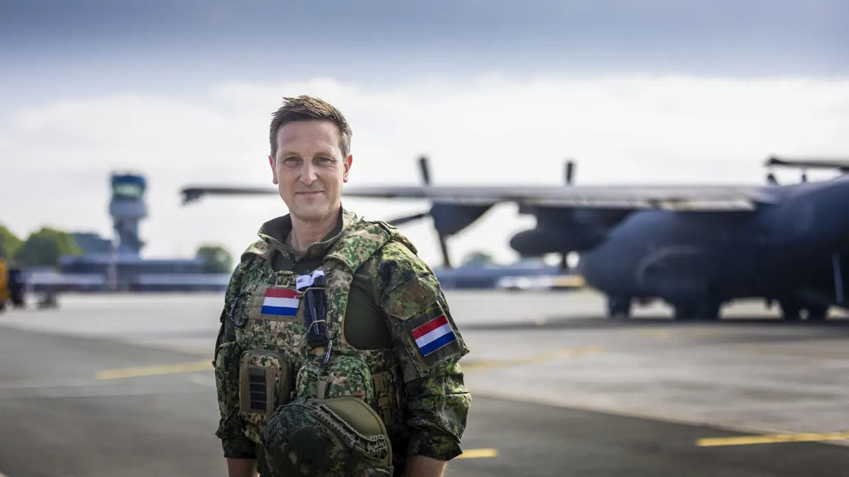 Militair met vest om staat op vliegveld met op de achtergrond een C-130 Hercules-transportvliegtuig.