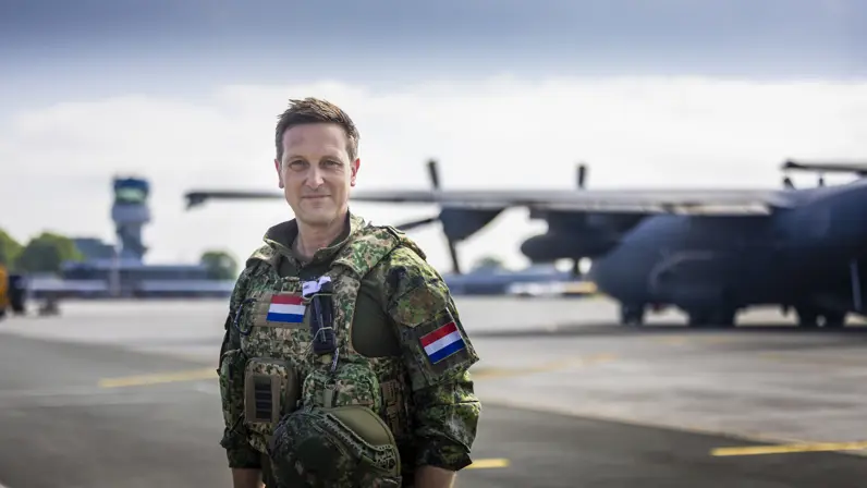Militair met vest om staat op vliegveld met op de achtergrond een C-130 Hercules-transportvliegtuig.