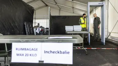 Militair in geel hesje geeft andere militair uitleg bij incheckbalie op tijdelijke vliegbasis.