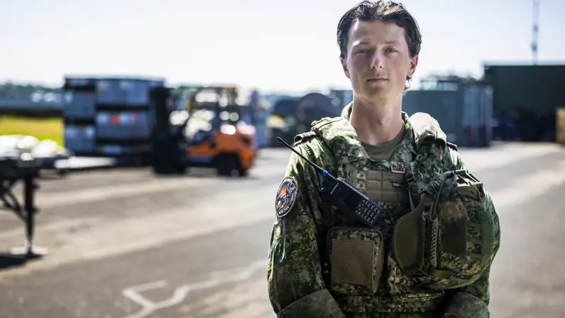 Militair met vest waaraan een helm hangt, staat op taxibaan vliegveld.